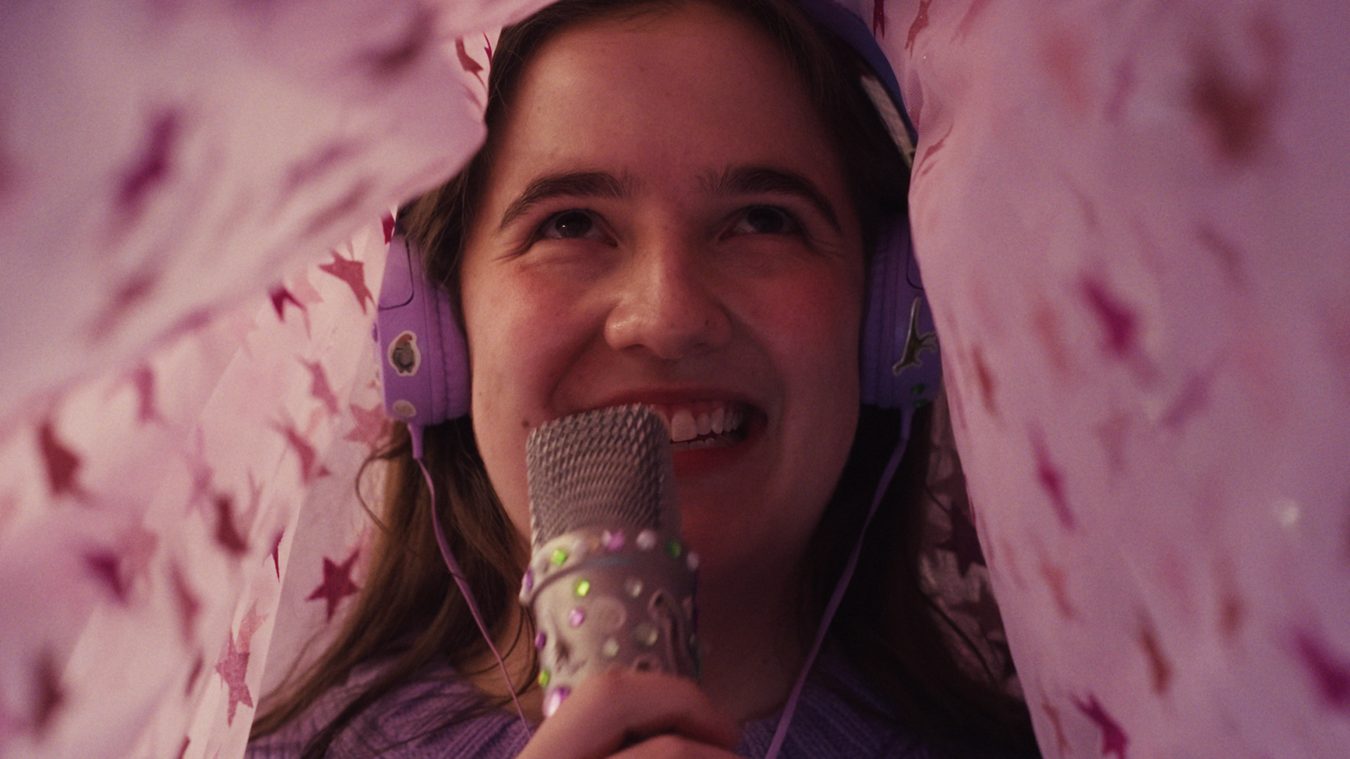 A smiling girl holding a microphone decorated with stickers sitting under a sheet covered in stars